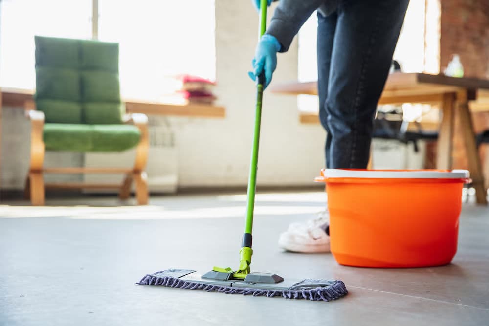 Person mopping a floor with a green mop and orange bucket in a well-lit room.